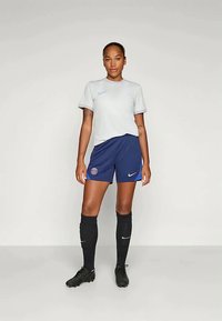 Naine seisab valgel taustal, seljas valge lühikese varrukaga spordipoliit, sinised PSG jalgpallilühikesed püksid, mustad põlvikud ja mustad nöörketsid.