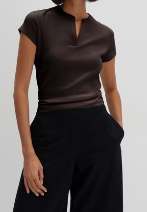 Nainen pukeutuneena istuvaan, lyhythihaiseen tummanruskeaan ribbineulepaitaan, jossa on pieni V-pääntie, ja löysiin mustiin korkeavyötäröisiin housuihin, käsi taskussa.