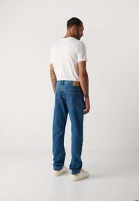 Mann med tatoveringer iført hvit t-skjorte, blå jeans og hvite joggesko stående og ser ned i et helt hvitt studio.