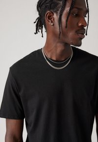 Juoda medvilninė marškinėlė su trumpomis rankovėmis ir apvalia iškirpte, pasižyminti lygiu paviršiumi ir sluoksniuotomis sidabrinėmis grandinėlėmis aplink kaklą.