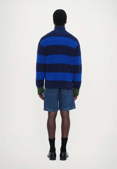 Blue and navy striped knit sweater with high collar, paired with denim shorts. Black socks and shoes complete the outfit. Back view shown.