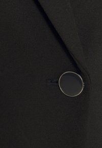 Blazer en tissu noir avec un grand bouton rond noir bordé d'argent métallique près d'une boutonnière.