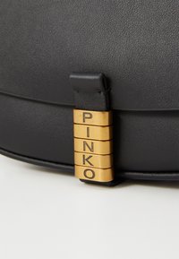 Melnas ādas soma ar gludu tekstūru. Ar zeltītu aparatūru, kurā ir iegravēti burti "PINKO". Kompakts, noapaļots dizains.