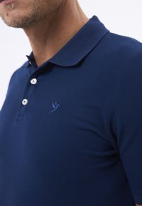 Tmavě modré polo tričko z texturované tkaniny s límečkem a třemi knoflíky, se vyšívaným logem na levé straně hrudníku.