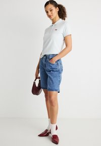 Νεαρή γυναίκα με σγουρά μαλλιά που φοράει ανοιχτό μπλε πόλο μπλούζα, μπλε τζιν σορτς, λευκές κάλτσες, μπορντό μυτερά παπούτσια και κρατάει μια μπορντό τσάντα.