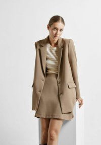 Blazer beige su misura con rever a punta e due tasche anteriori, abbinato a una gonna coordinata. Top fitted con texture e righe chiare.