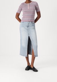 Personne portant un haut à rayures à manches courtes, une jupe midi en denim bleu clair avec une fente avant, et des chaussures noires sans lacets sur un fond blanc.