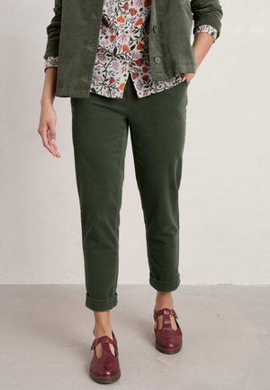 Persoon met olijfgroene opgerolde broek, bloes met bloemenprint, bijpassend jasje en bordeauxrode gesloten T-strap schoenen, staand op een lichte vloer.