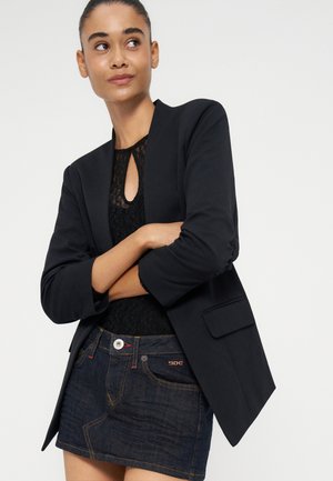 Frau trägt einen schwarzen Blazer über einem schwarzen Spitzenoberteil und einem dunklen Denim-Minirock, steht mit verschränkten Armen und schaut leicht zur Seite.