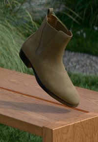 Brown suede Chelsea boot with a sleek silhouette, elastic side panels, and a smooth black sole, presented on a wooden surface.