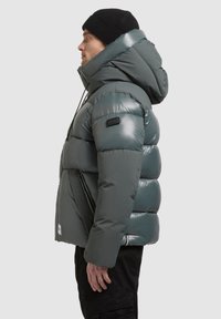 Męska kurtka puchowa w ciemnozielonym kolorze, błyszczący materiał, kaptur, pikowane sekcje, z czarną naszywką z logo i sznureczkami do regulacji.