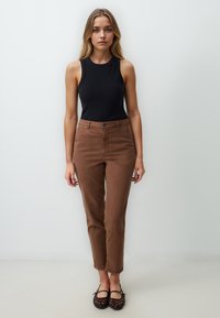 Donna con lunghi capelli biondi, che indossa un top nero senza maniche, pantaloni marroni a vita alta e ballerine nere intrecciate, in piedi contro un muro bianco.