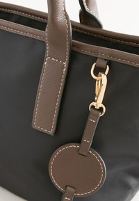 Sac cabas noir avec poignées en cuir marron, étiquette ronde en cuir cousue et fermoir doré sur la sangle latérale.