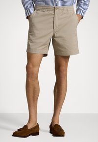 Pantaloni corti in cotone beige con elastico in vita e tasche frontali, abbinati a scarpe slip-on in camoscio marrone. Orlo ben rifinito e design semplice.