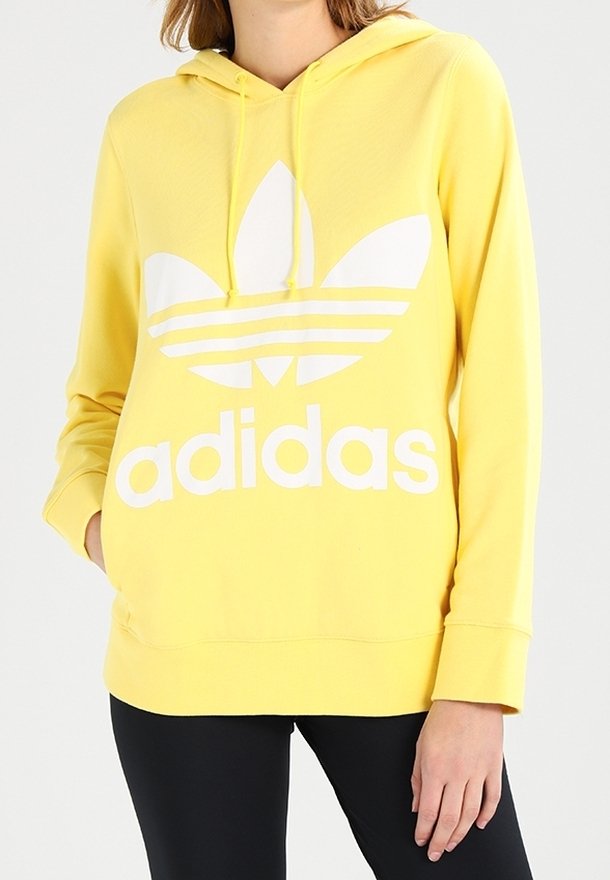 Gul huvtröja med en stor vit Adidas-logga. Den har långa ärmar, en framficka och ett slätt, strukturerat tyg.