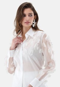 Weiße Bluse mit transparentem Stoff und floraler Stickerei auf den Ärmeln, durchgehend zugeknöpft und mit klassischem Kragen. Glatte Textur mit dezentem Design.