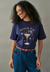 Marine crew-neck t-shirt med et farverigt "Tinsel Tini" grafik af et cocktailglas med stjerne- og fyrværkeri-accenter. Båret med blå jeans og et koralfarvet bælte.