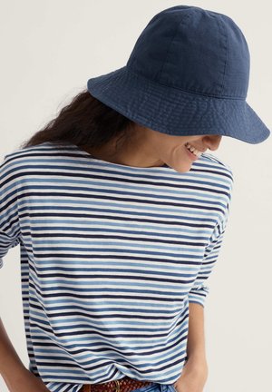 Naeratav naine, kandes tumelõhnav sinist tilgakübarat ja sinise, valge ning musta triibulise pika varrukaga särki, käed taskutes tavalise tausta ees.