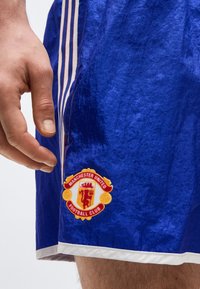 Main à côté d’un short de sport bleu avec le logo du Manchester United Football Club sur la jambe gauche.