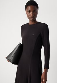 Melna garās piedurknes kleita ar smalku logo uz krūtīm, apvienota ar struktūrētu melnu ādas rokassomu. Gluda auduma un pielāgota piegriezuma.