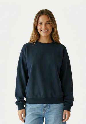 Jonge vrouw glimlachend, gekleed in een effen donkerblauw crewneck-sweatshirt en lichtblauwe jeans, staand tegen een witte achtergrond.
