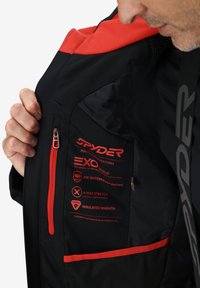 Schwarze Jacke mit roten Akzenten und Reißverschlusstasche. Enthält Leistungslabel, die Materialien, Wasserdichtigkeitsbewertung und Isolierung detaillieren.