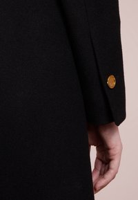 Primo piano di un cappotto di lana nera, che mostra una texture liscia, un design su misura e un dettaglio con bottone dorato sulla manica.