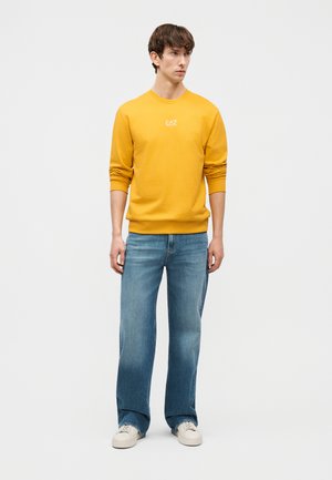 Noor mees seisab, kandes sinepikollast EA7 dressipluusi, siniseid teksaseid ja valgeid tenniseid, tavalise heletaustaga.