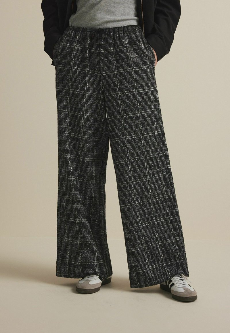 Pantalon à jambes larges avec un motif à carreaux noir et blanc texturé ; il est doté d'une ceinture élastique à cordon de serrage et de poches avant.