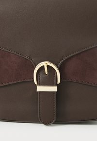 Sac en cuir marron avec un accent en daim bordeaux foncé, une boucle en or et une texture lisse avec des détails cousus.