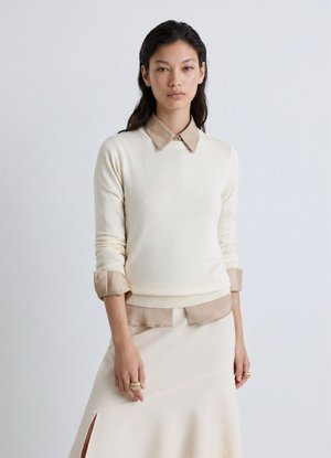 Donna con capelli lunghi e scuri che indossa un maglione crema sopra una camicia beige con colletto e una gonna crema abbinata con uno spacco laterale, in piedi davanti a uno sfondo semplice.