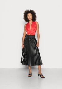 Top halter de seda vermelha com decote em V profundo, combinado com uma saia midi plissada preta. Os acessórios incluem uma pequena bolsa preta e sapatos de salto aberto.