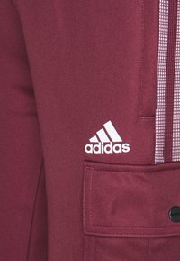 Bordeauxkleurige sportbroek met een gladde textuur, voorzien van het witte adidas logo en verticale strepenaccenten. Inclusief een detail van een zijvak.