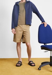 Marinvuxen hoodie i navy över en olivfärgad t-shirt, khakishorts och svarta sandaler. Står bredvid en blå kontorsstol på ett gult golv.
