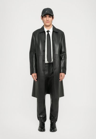 Man wearing black leather long coat, black leather pants, black leather shoes, white shirt, black tie, and black cap standing against white background.