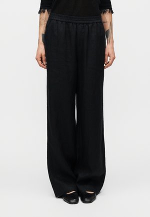Μαύρο λινό παντελόνι φαρδιάς γραμμής με ελαστική μέση, με λεία υφή και άνετη εφαρμογή, σε συνδυασμό με μαύρα παπούτσια.