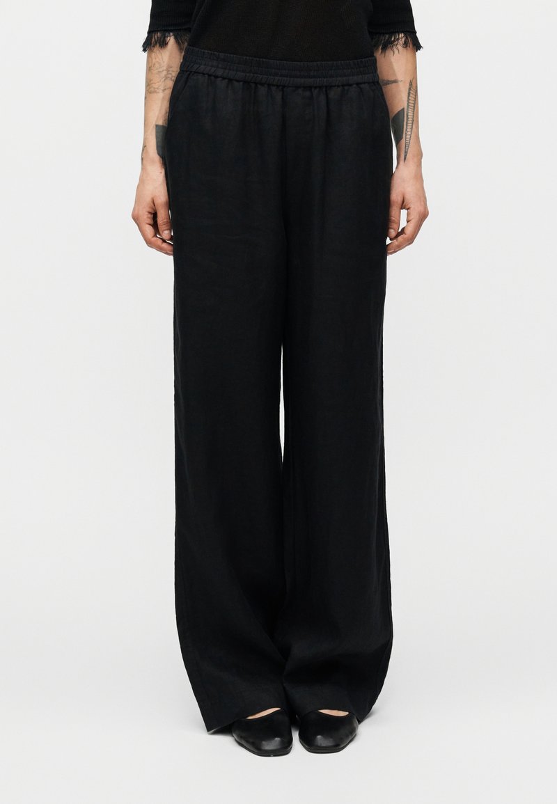 Μαύρο λινό παντελόνι φαρδιάς γραμμής με ελαστική μέση, με λεία υφή και άνετη εφαρμογή, σε συνδυασμό με μαύρα παπούτσια.