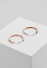 Brincos de aro em rosa dourado com um design suave e arredondado e uma ligeira abertura. Exibidos sobre um pedestal geométrico branco.