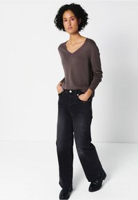 Szary sweter z dekoltem w serek połączony z luźnymi, czarnymi jeansami z wysokim stanem. Model stoi z rękami w kieszeniach, nosząc czarne botki za kostkę.