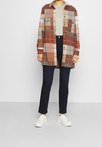 Camisa de franela a cuadros en tonos marrón, rojo y gris, con dos bolsillos en el pecho y cierre frontal de botones, usada sobre una blusa verde clara y jeans oscuros.