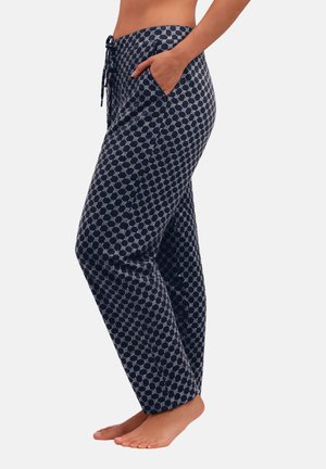 Kvinde iført løsthængende marineblå bukser med mønster og sidelommer, stående barfodet mod en ensartet lys baggrund.