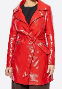 Femme portant une veste en simili cuir rouge brillant ceinturée avec des poches zippées, sur un haut noir et un pantalon marron.