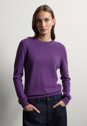 Giovane donna con capelli castani che indossa un maglione viola a maniche lunghe e jeans scuri, in piedi davanti a uno sfondo chiaro e uniforme con le mani in tasca.