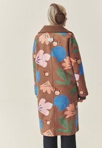 Brązowy płaszcz z kwiatowym wzorem w kolorach różowym, niebieskim i zielonym, ozdobiony twarzami niedźwiedzi oraz teksturowanym kołnierzem. Ciepłe nauszniki w kolorze brązowym.
