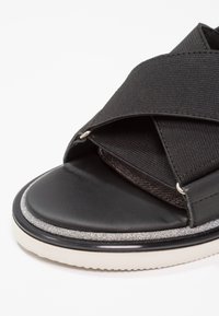 Sandale noire avec des brides croisées en tissu texturé, une semelle intérieure en cuir lisse et une semelle en caoutchouc avec un accent métallique.