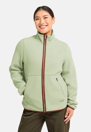 Vrouw die lacht en een lichtgroen fleeces jas met bruine rits en bies draagt, handen in de zakken, tegen een effen witte achtergrond.