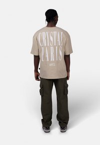 Beżowy oversize'owy t-shirt z białym napisem "CRYSTAL PARIS" oraz "DEPT.75" z tyłu. Noszony z oliwkowymi spodniami cargo i szarymi butami.
