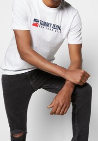 Vit bomull t-shirt med "TOMMY JEANS NEW YORK CITY"-logotyp i marinblått och rött. Stylar med smala svarta jeans med rivna knän.