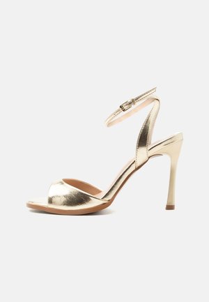 Guld højhælet sandal med ankelrem og spænde, åben tå og slank stiletthæl på en ensartet lys baggrund.