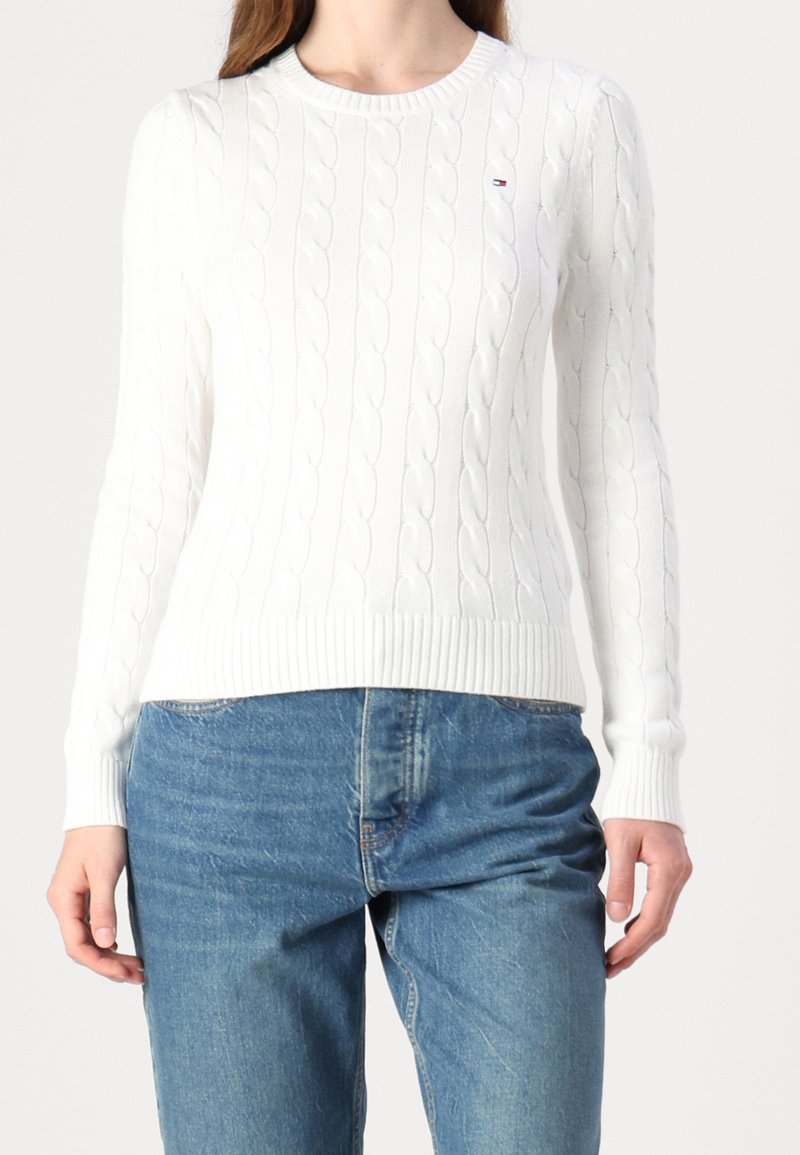 Biały sweter w splot warkoczowy z prążkowanym dekoltem i brzegiem. Noszony z jasnoniebieskimi dżinsami, zawiera mały akcent logo na klatce piersiowej.